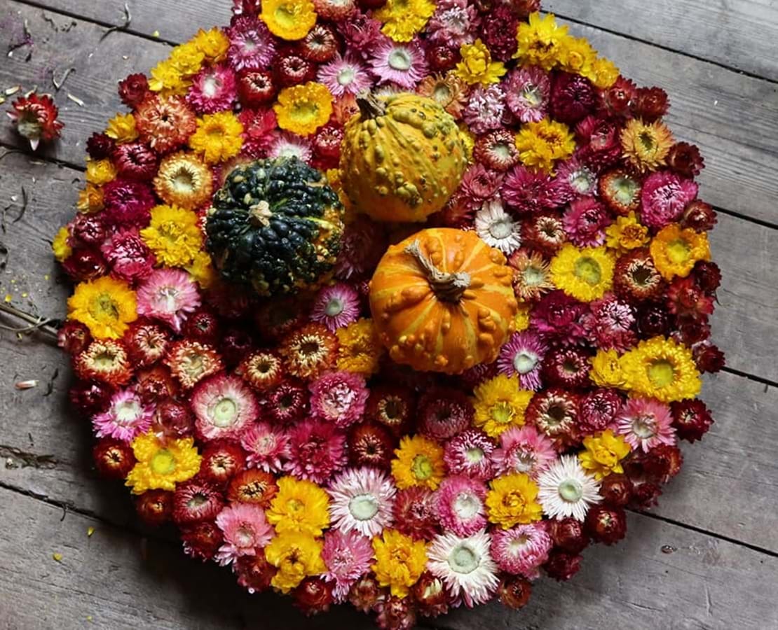 Rund form en tæt bundet blomster i gule, lilla og røde efterårsfarver. På toppen ligger tre græskar i grøn, gul og orange farve.