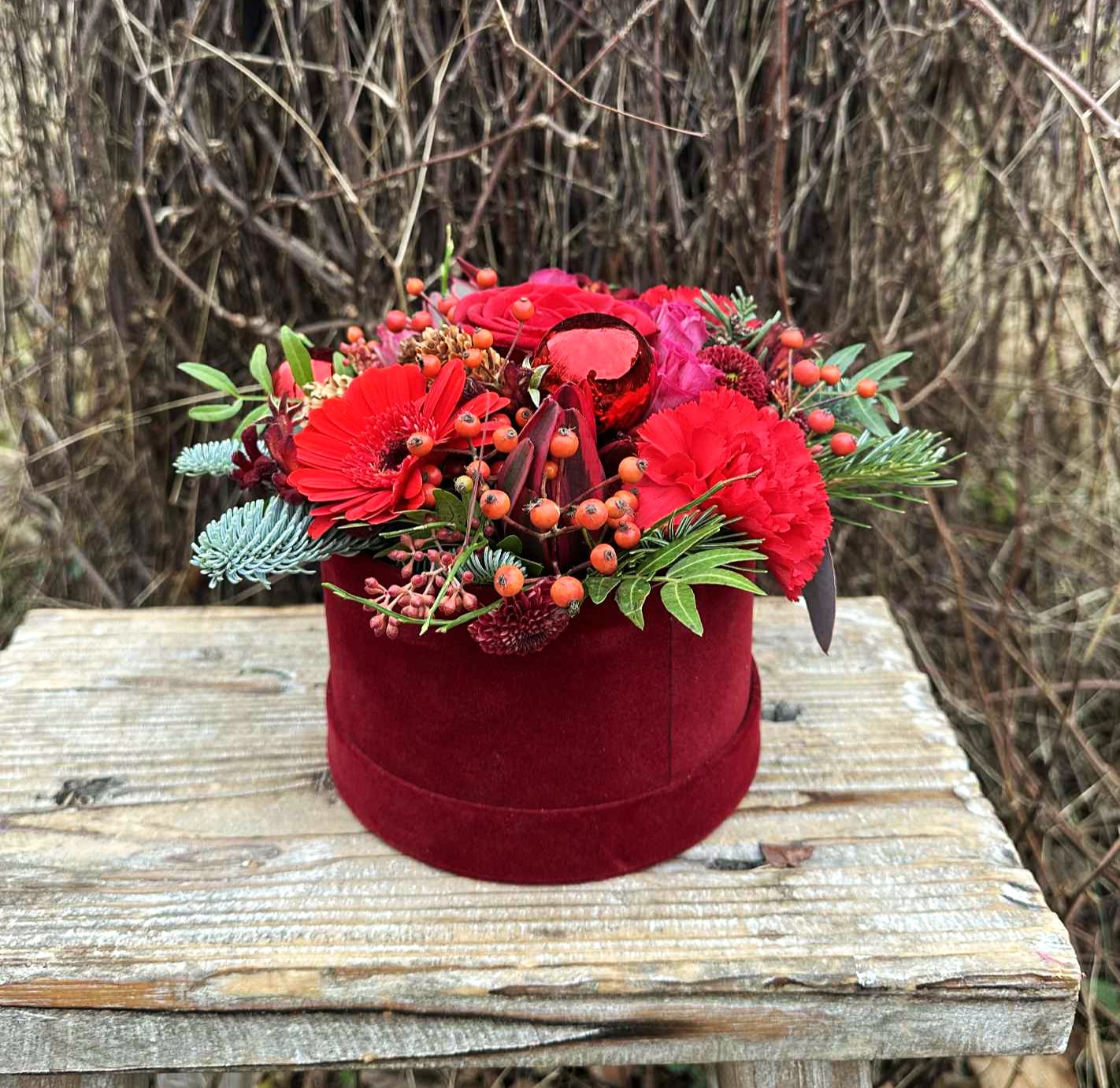 Rød julehatteæske med roser, gran, bærgrene og røde gerbera. 
